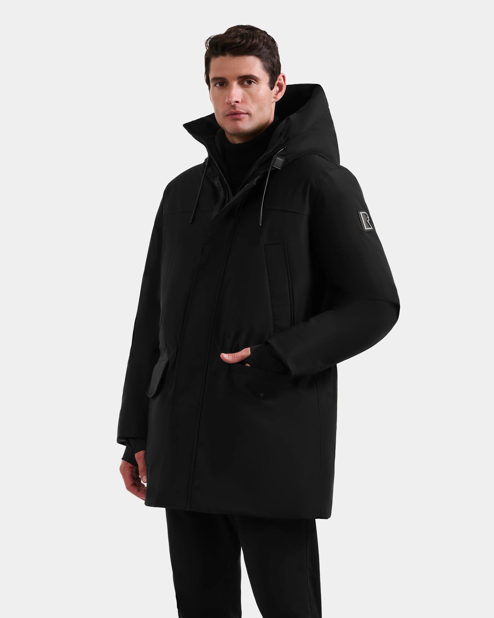 A man in the Rudsak MITCHELL - M125513 Black long men’s down parka with high collar and hood stands against a white background, one hand by his side, the other near the zipper, looking forward.