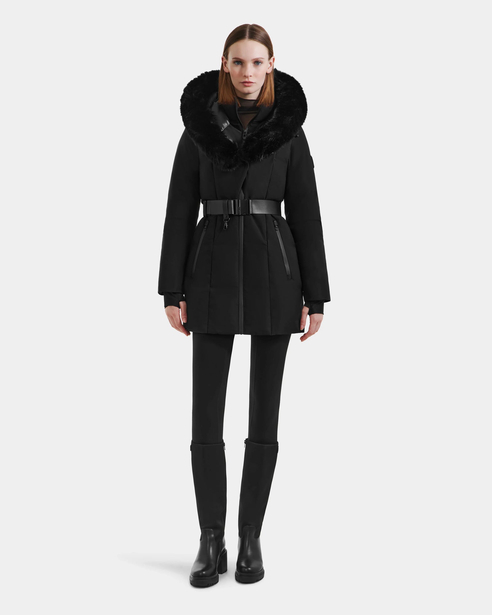 A woman faces forward against a white background, wearing the Rudsak MODA M - L125555 Black down puffer coat with a vegan leather belt and faux fur hood, paired with black leggings and boots.
