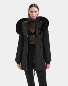 A woman faces forward against a plain white background wearing the Rudsak MODA M - L125555 Black winter coat with a large, fluffy fur-lined hood, paired with a sheer black top and black leggings.
