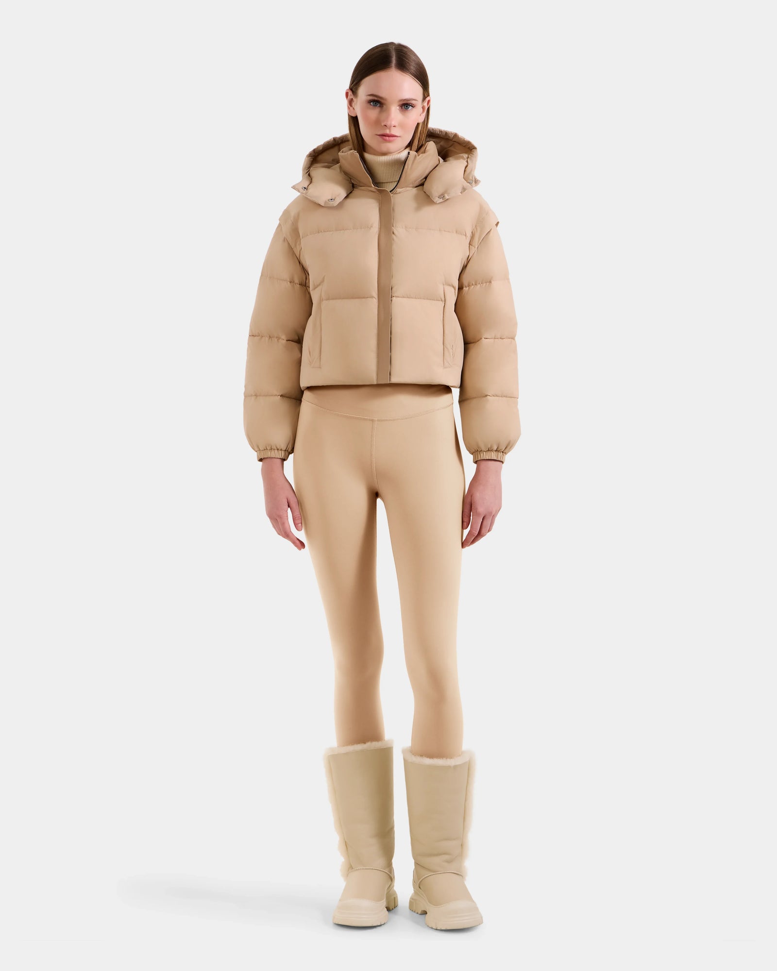 A woman faces forward against a plain white background, wearing the Rudsak OPRAH - L125546 Vanilla beige cropped down puffer, paired with matching beige leggings and fur-lined boots.