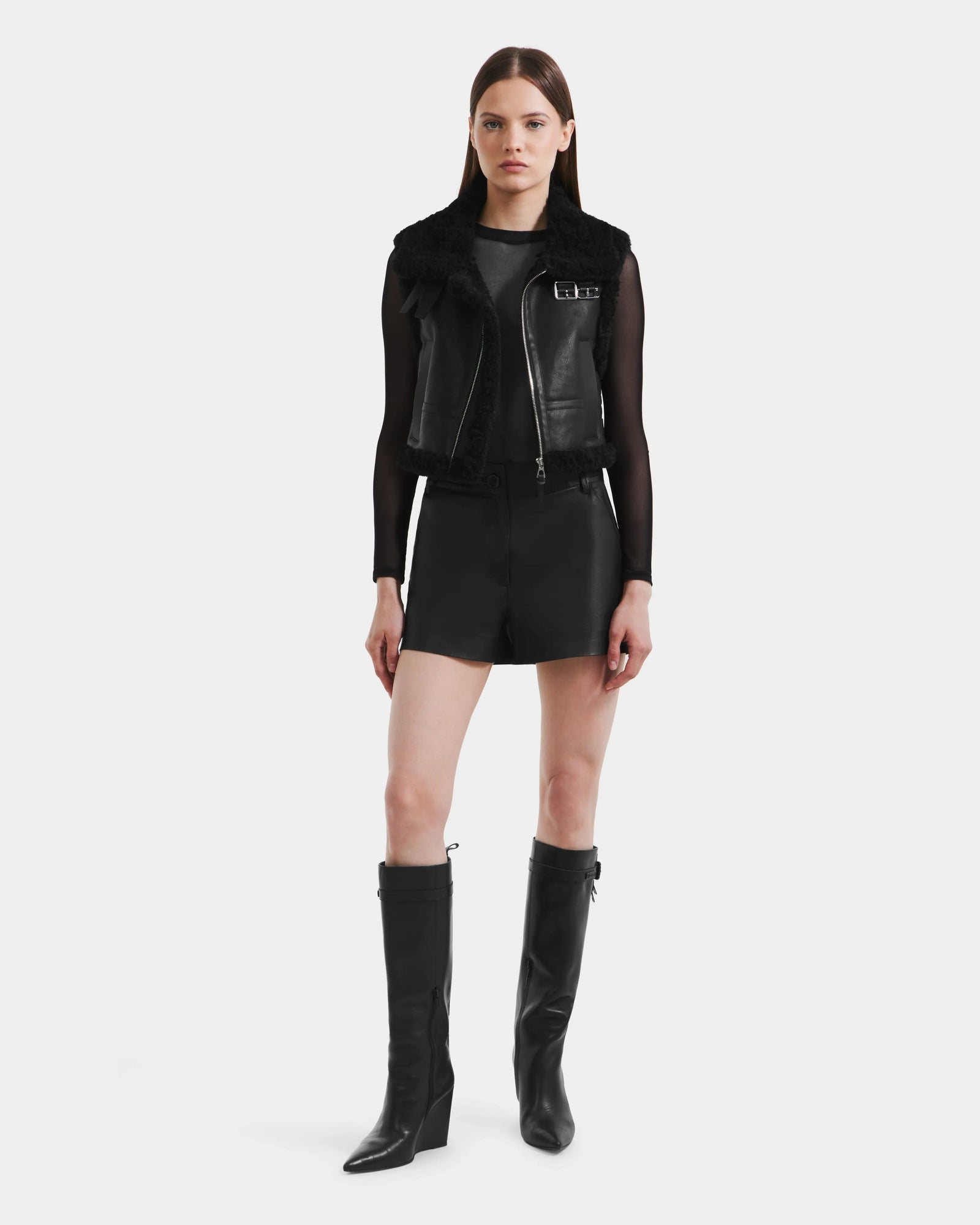 A woman faces forward in a Rudsak PAISLEY - L125516 Black vegan leather vest with faux shearling, sheer long-sleeve top, black shorts, and knee-high pointed-toe boots against a plain white background.