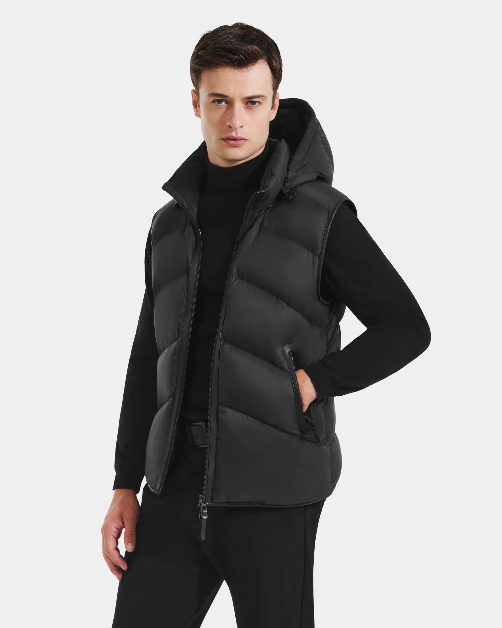 A man models the Rudsak RICHARDSON - M125527 Black water-repellent men’s hooded vest over a black turtleneck and pants, highlighting its lightweight warmth and modern style against a plain white background.