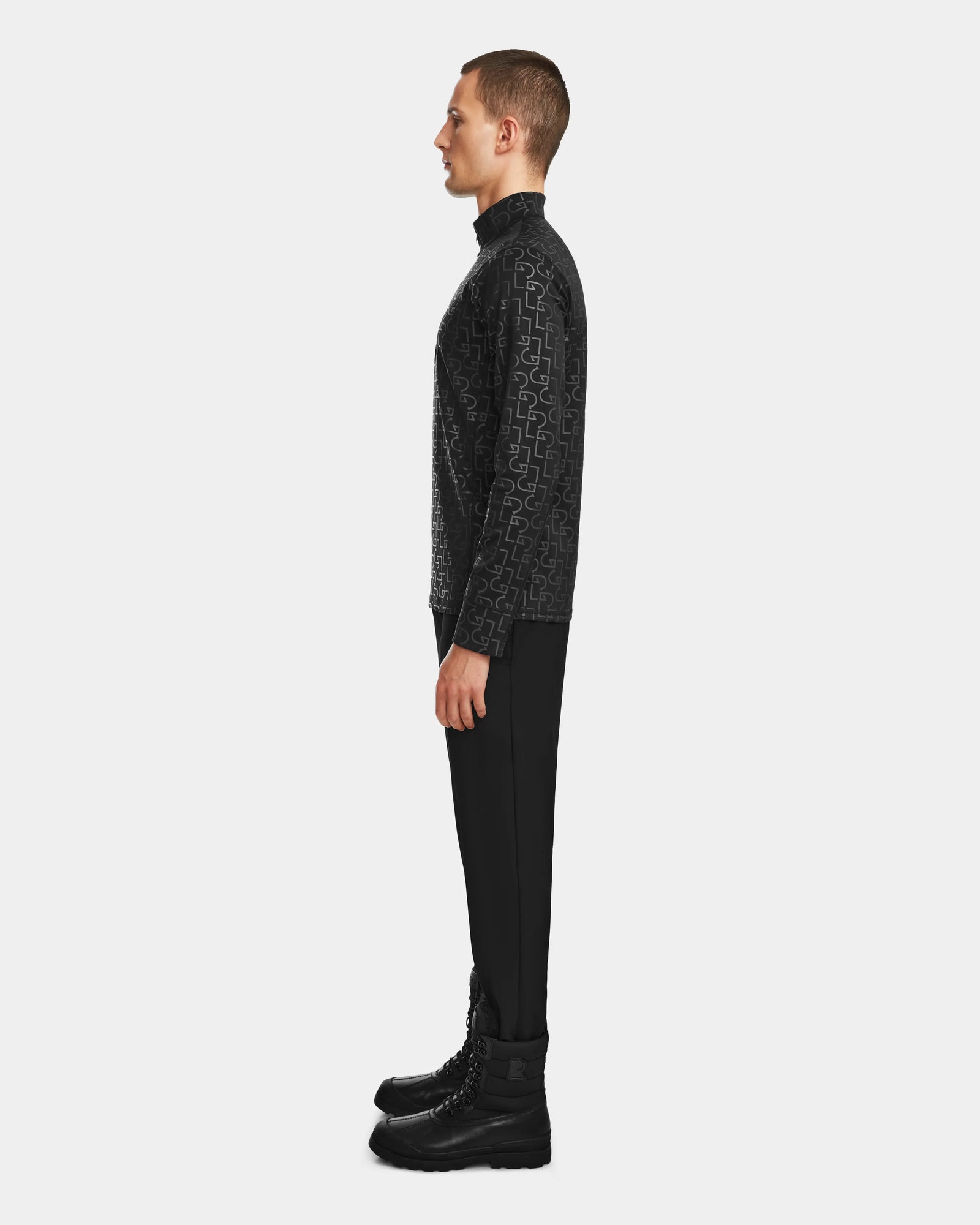 A man stands in profile against a plain background, wearing the Rudsak ROMEO - 6524525 Black stretch active top—a black patterned, long-sleeve, mock-neck zip-up—paired with black pants and lace-up boots.