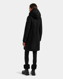 A woman with straight brown hair faces away, wearing the SARINA - L125550 Black parka by Rudsak, paired with black leggings and black fur-cuff boots. The background is plain light gray.