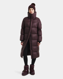 A person wearing the Rudsak SASHA - 8125686 Bordeaux long down puffer coat, matching boots, black leggings, and a maroon knit beanie stands against a plain white background.