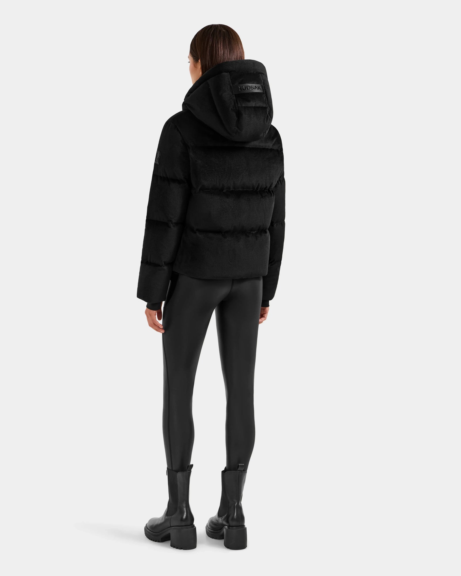 A person with straight brown hair, seen from the back, models the Rudsak STEFANIE V - L125584 Black down puffer jacket with a hood, shiny black leggings, and black ankle boots against a plain light background.