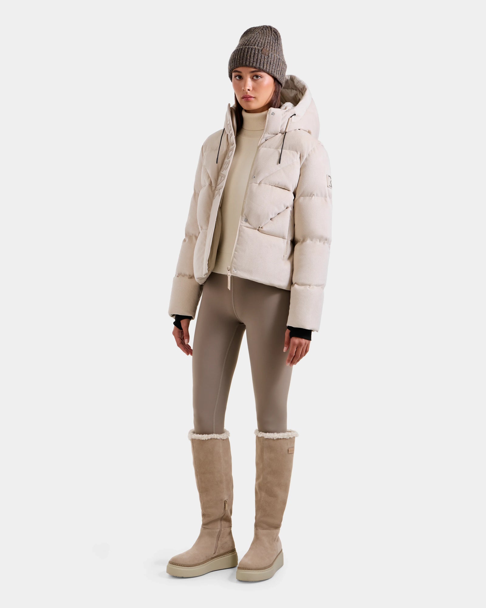 A woman stands against a white background wearing the Rudsak STEFANIE V - L125584 Vanilla puffer jacket, styled with a beige turtleneck, taupe leggings, knee-high fur-trimmed boots, and a gray knit beanie.
