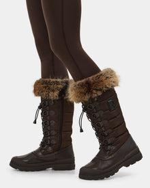 A person showcases the Rudsak WINDY - L224507 Espresso women’s winter boots with lace-up fronts, side zippers, and faux fur trim, styled with dark leggings against a plain white background.