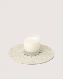 A white candle in a frosted glass holder by Graf Lantz rests on a round, textured beige coaster against a plain light background.