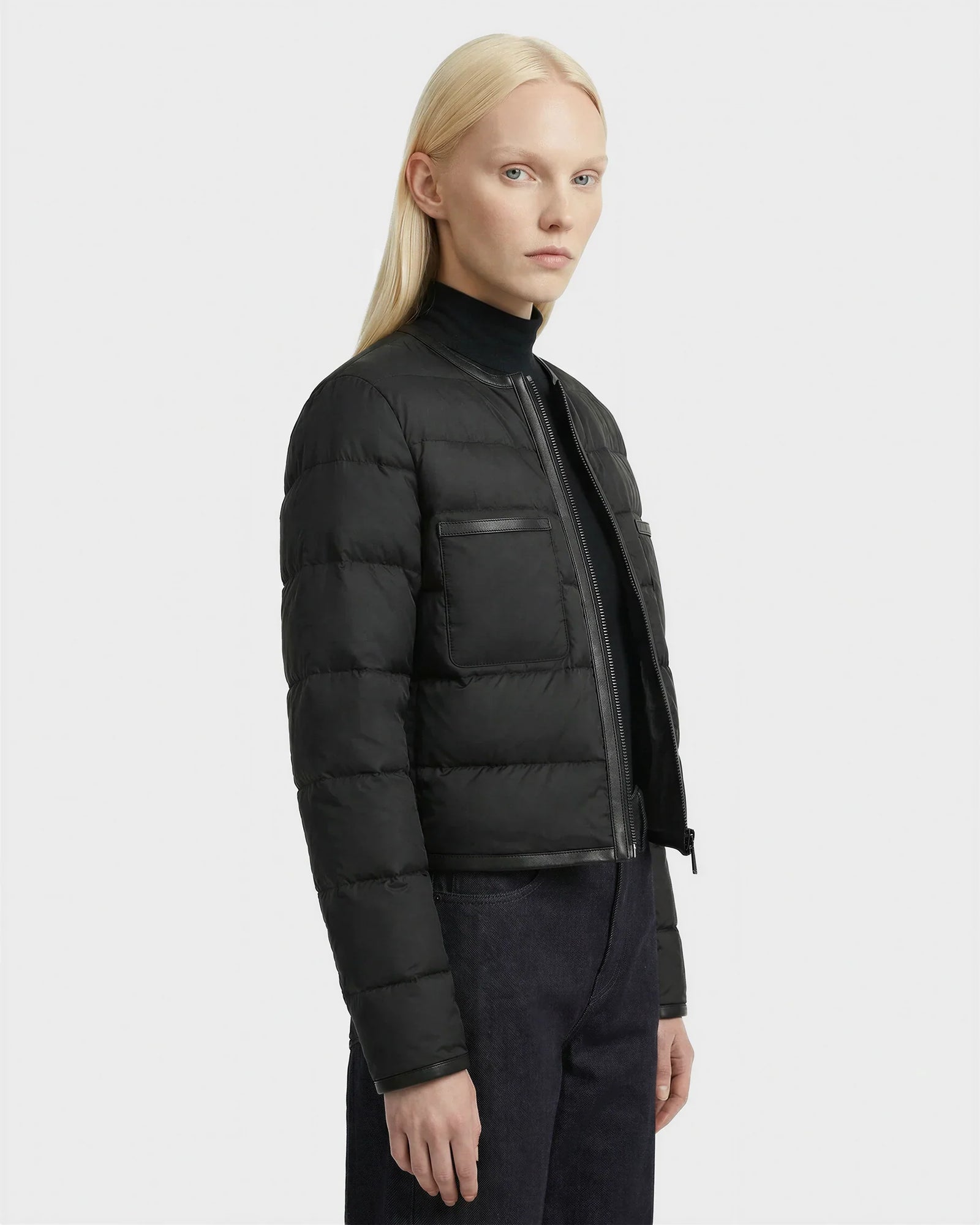 A person with long blond hair wears the Rudsak MARKLE - 8125033 Black women’s cropped down jacket with vegan leather trim, a black turtleneck, and dark pants, standing sideways against a plain white background.