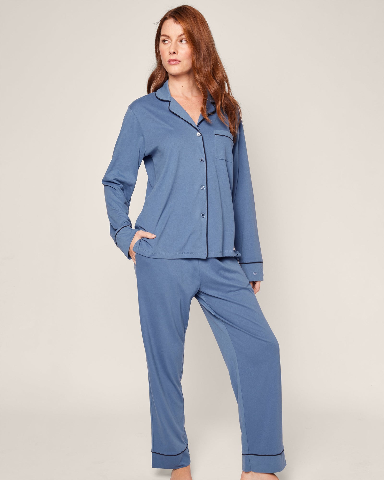 A person stands against a light-colored background wearing Petite Plume’s Womens Pima Pajama Set in Mystique Blue, made from luxurious Peruvian Pima cotton. The set includes a long-sleeved button-up shirt and pants, with one hand tucked in their pocket.