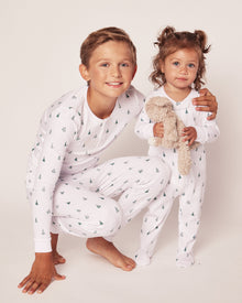 Two smiling children are photographed wearing Petite Plumes Kids Pima Snug Fit Pajama Set in Evergreen Trees. The older child wears a long sleeve top and pants while crouched beside the younger one holding a plush toy, set against a plain, light-colored background.