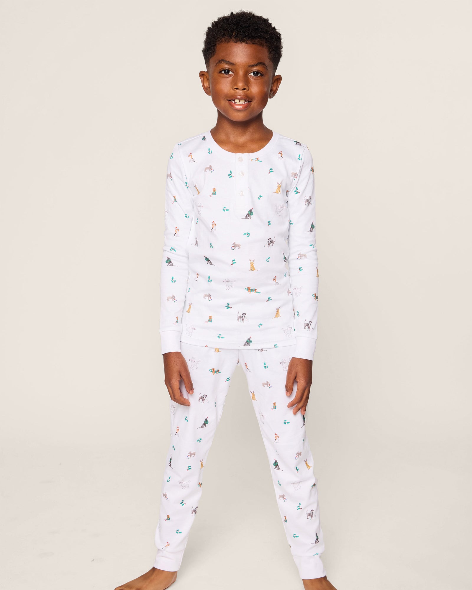 A young boy smiles, wearing the Petite Plume Kids Pima Snug Fit Pajama Set in Jingle Paws. This luxurious sleepwear features colorful small animal prints and provides comfort with its long sleeve top and pants as he stands barefoot against a plain background.