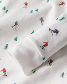 A close-up of Petite Plumes Apres Ski Kids Pima Snug Fit Pajama Set showcases skiers in vibrant attire, moving in various directions on soft and cozy white fabric, dotted with small green pine trees—ideal for a snug long sleeve top or pant set.