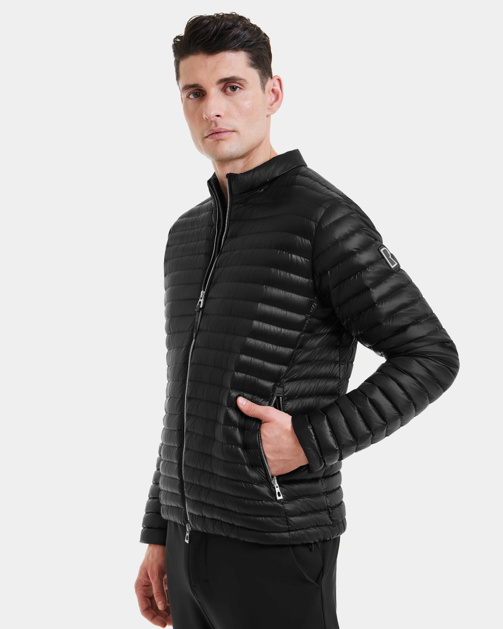 A man wears the Rudsak REYMOND - M125013 Black, a quilted mens down jacket made from recycled nylon with RDS-certified down, standing with his right hand in the pocket against a plain white background.