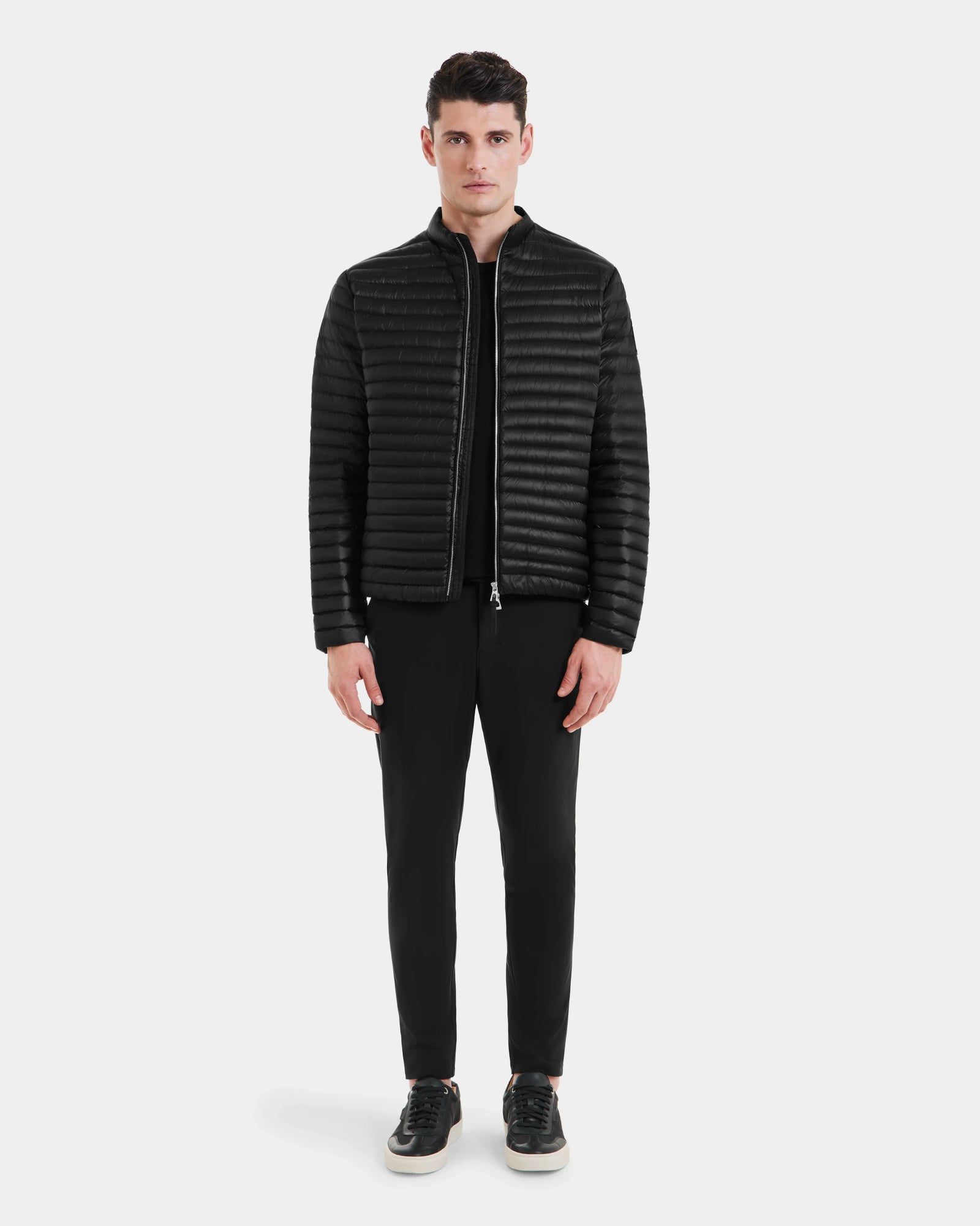 A man faces forward wearing the Rudsak REYMOND - M125013 Black men’s down jacket, quilted and made from recycled nylon, paired with black pants and black sneakers with white soles against a plain light gray background.