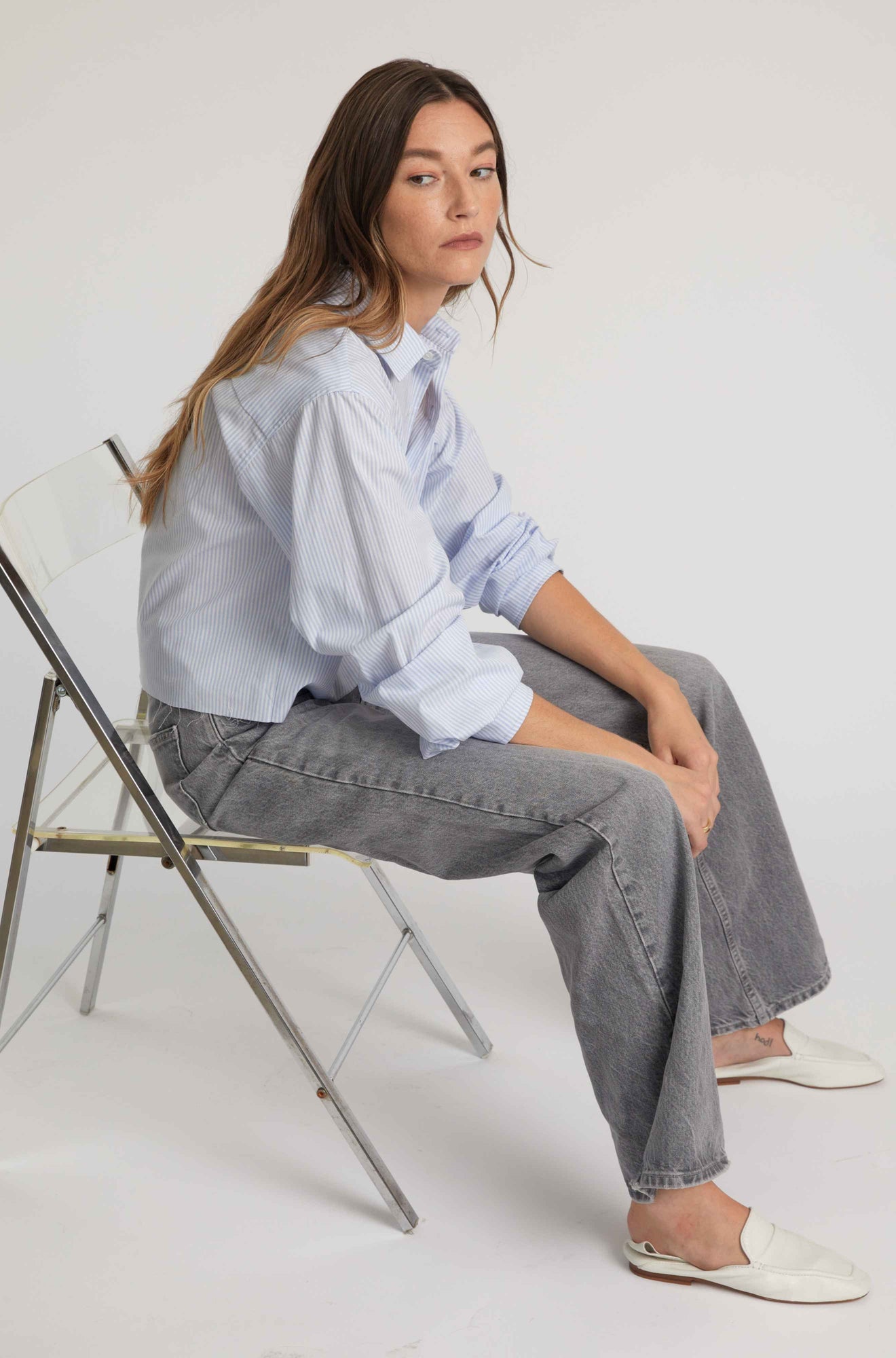 Woman sitting on a chair wearing a light blue shirt and gray pants against a white background
