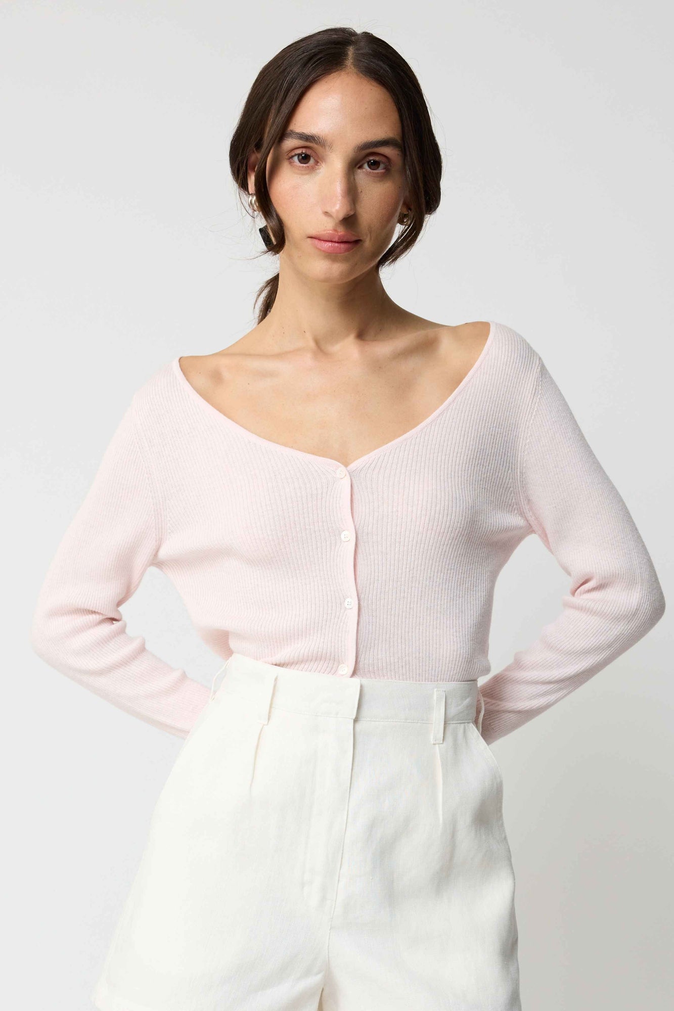 Woman wearing a light pink long-sleeve top and white pants on a plain background