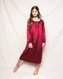 A girl wearing Petite Plumes Girls Silk Delphine Nightgown in Bordeaux Polka Dot stands barefoot on a beige fabric background. Her long hair falls over her shoulders as she gazes downward, with one leg slightly bent forward.