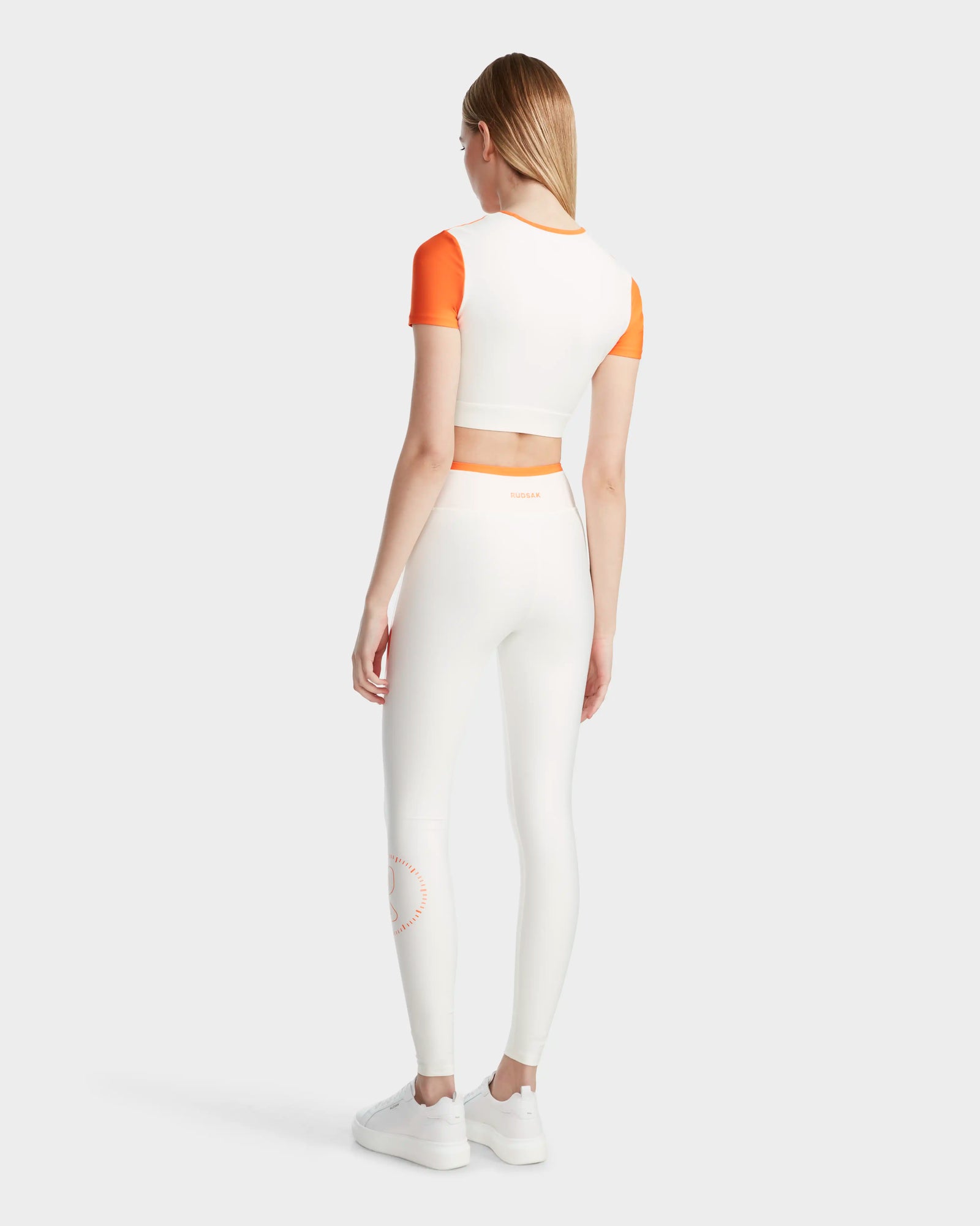 A woman with straight blonde hair wears the Rudsak LILIANE - 8524018 Eggshell womens fitted crop top and high-waisted leggings, made from high-performance stretch fabric. She faces away from the camera in white sneakers on a plain white background.