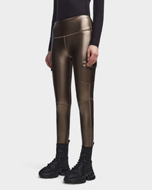 A person wearing Rudsaks SIEN P - 8523027 METALLIC CHOCOLATE stretch vegan leather cargo leggings with side pockets, a black long-sleeve top, and black high-top sneakers, standing against a plain white background.