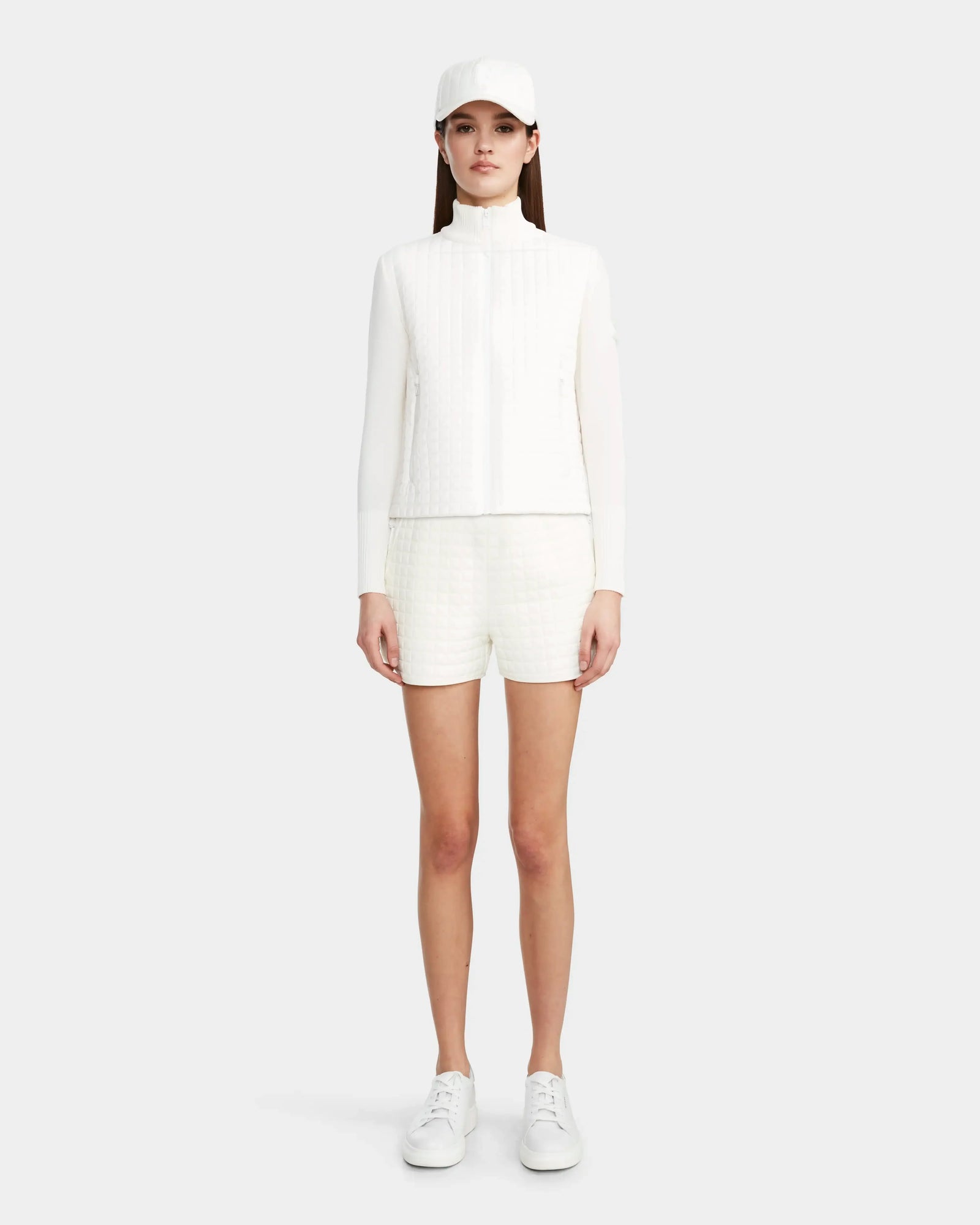 A woman faces forward wearing the Rudsak LOUISE - 8124046 Eggshell lightweight quilted jacket with matching white shorts, styled with a white baseball cap and sneakers against a plain light background.