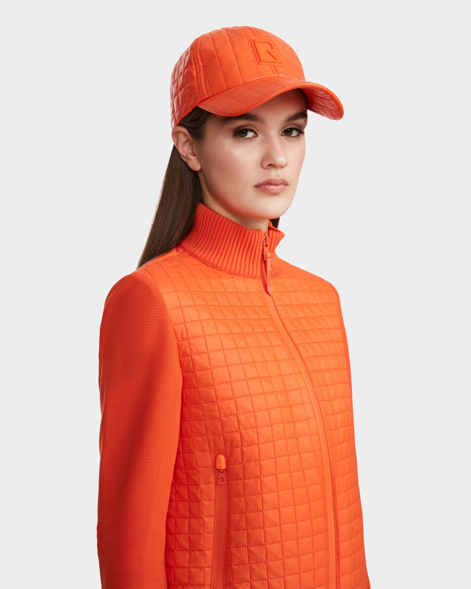 A woman in the Rudsak LOUISE - 8124046 Orange Sorbet lightweight quilted jacket and matching quilted baseball cap looks at the camera with a neutral expression against a light gray background.