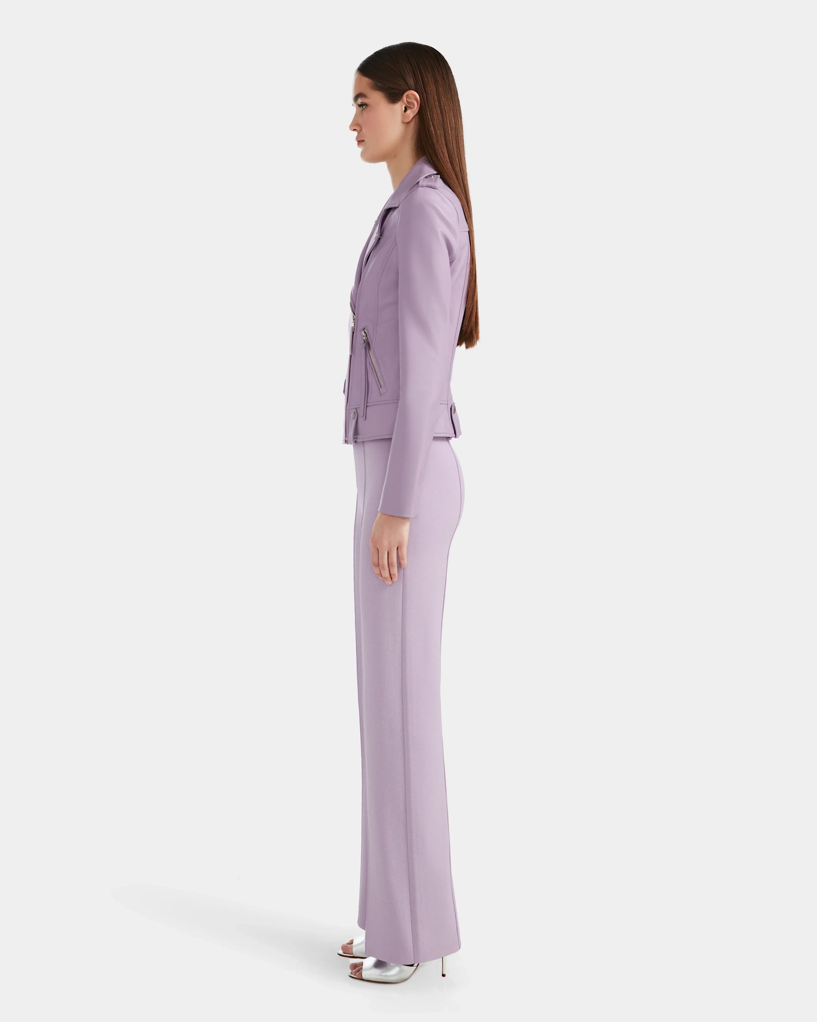 A woman in profile models the Rudsak MERGO - 8124034 Lilac Foam fitted pantsuit, inspired by a womens biker jacket with zip accents, paired with silver high heels against a plain white background.