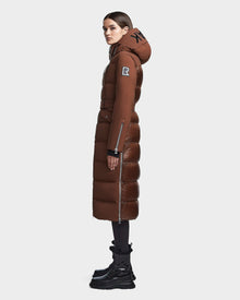 A woman stands in profile against a white background wearing the RUDSAK CALI - 8122502 COHIBA, a long brown women’s winter puffer coat with hood, RDS-certified down, logo on hood and sleeve, side zippers, black leggings, and lace-up boots.