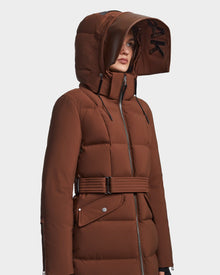 A person wears the RUDSAK CALI - 8122502 COHIBA, a brown belted womens winter down puffer coat with a large, structured hood featuring black lettering on the inner brim, zip front, and snap pockets, looking slightly to the side.