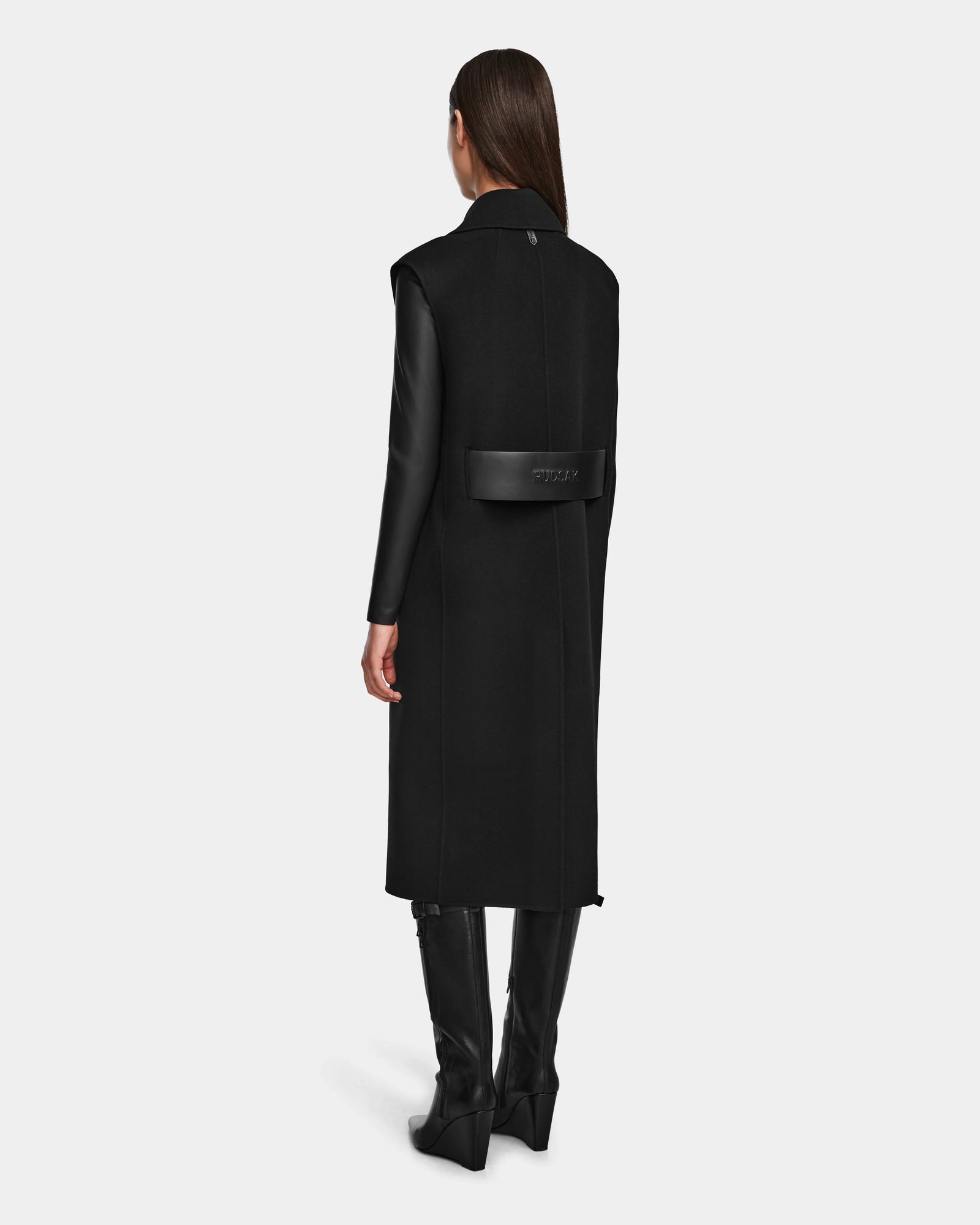 A woman faces away, wearing the Rudsak NOURA - 8124698 black knee-length wool vest with leather sleeves, black knee-high boots, and a black Givenchy belt bag. The background is plain white.
