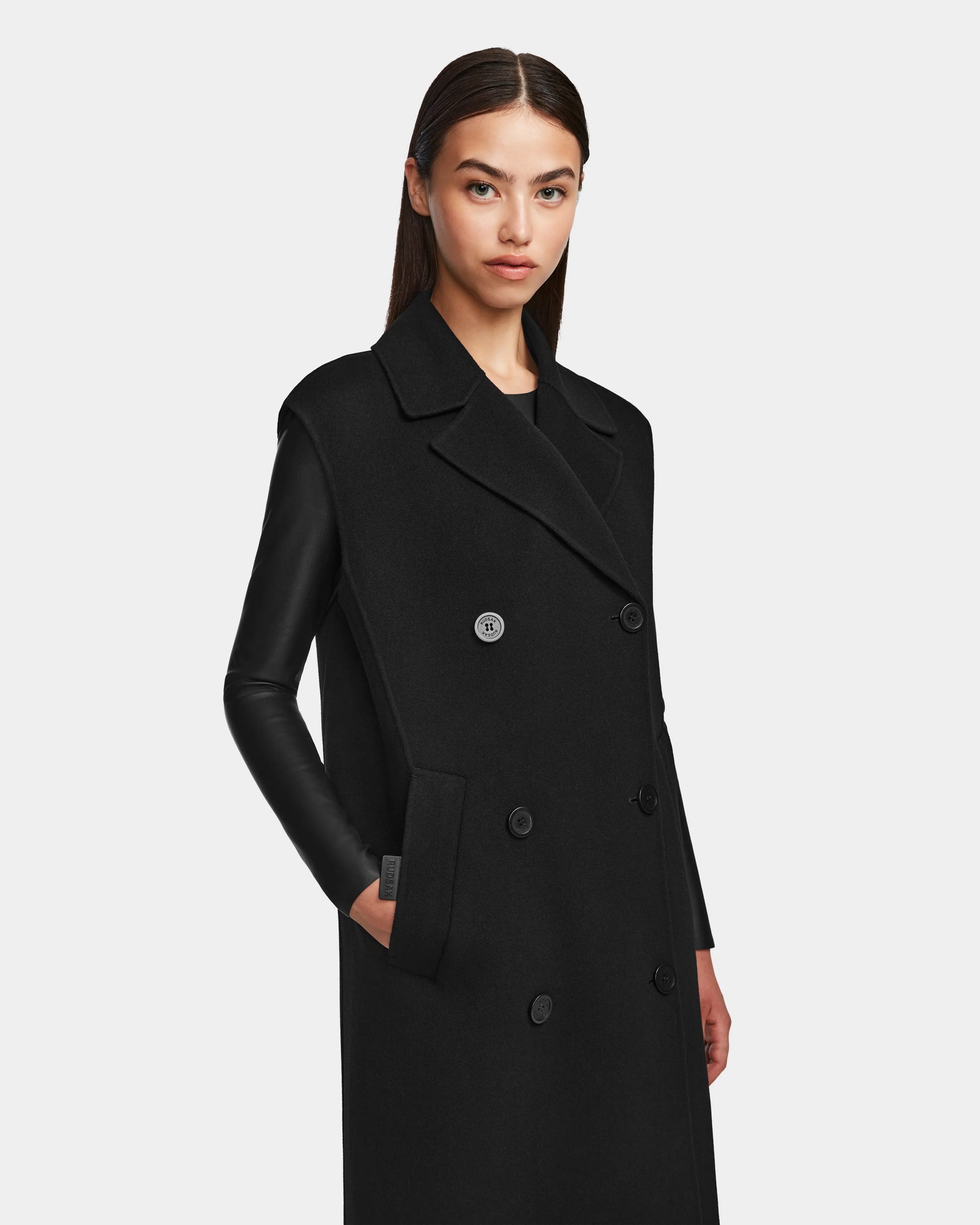 A woman with straight, dark hair models the Rudsak NOURA - 8124698 Black knee-length vest with leather sleeves, standing with one hand in her pocket against a light gray background.