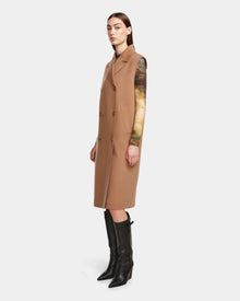 A woman stands sideways against a white background wearing the Rudsak NOURA - 8124698 Camel double-breasted vest styled as a long coat over a printed top and knee-high black boots.