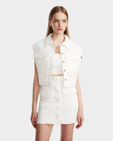 A woman with straight blonde hair wears the Rudsak SOLSA - 8124048 Eggshell womens cropped quilted vest over a white crop top and matching skirt, standing against a light gray background and looking slightly to her left.