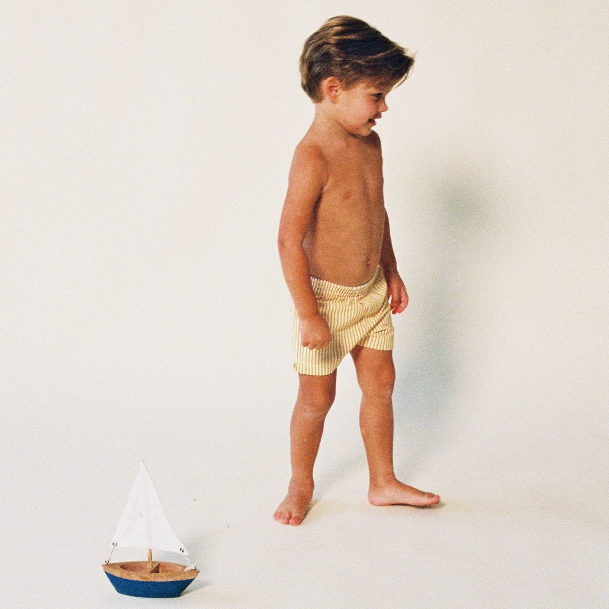 Child wearing yellow and white striped swim trunks, standing in profile against a neutral background.