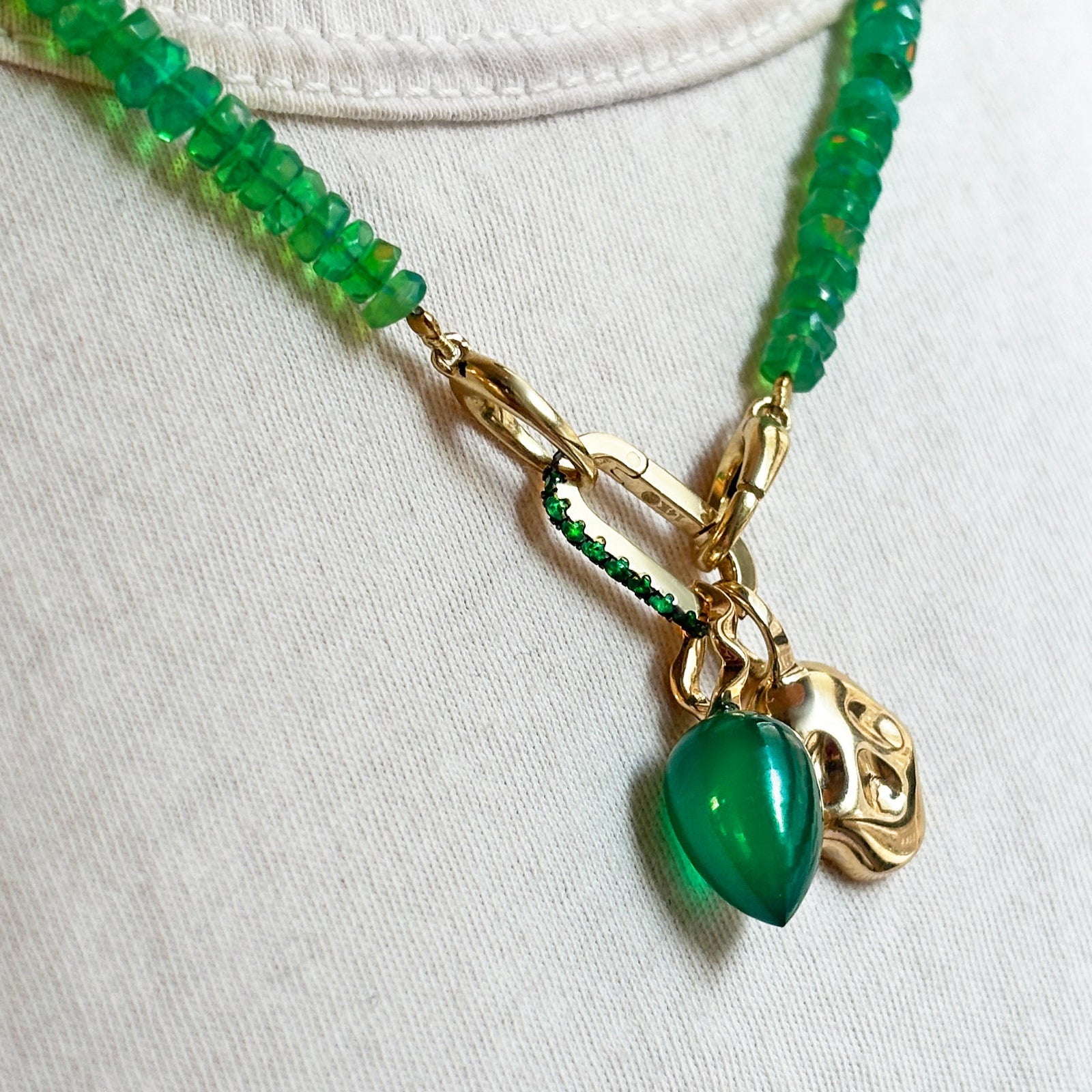 Green faceted opal necklace with molten skull charm, small emerald charm lock, and green onyx drop charm on a white tee.