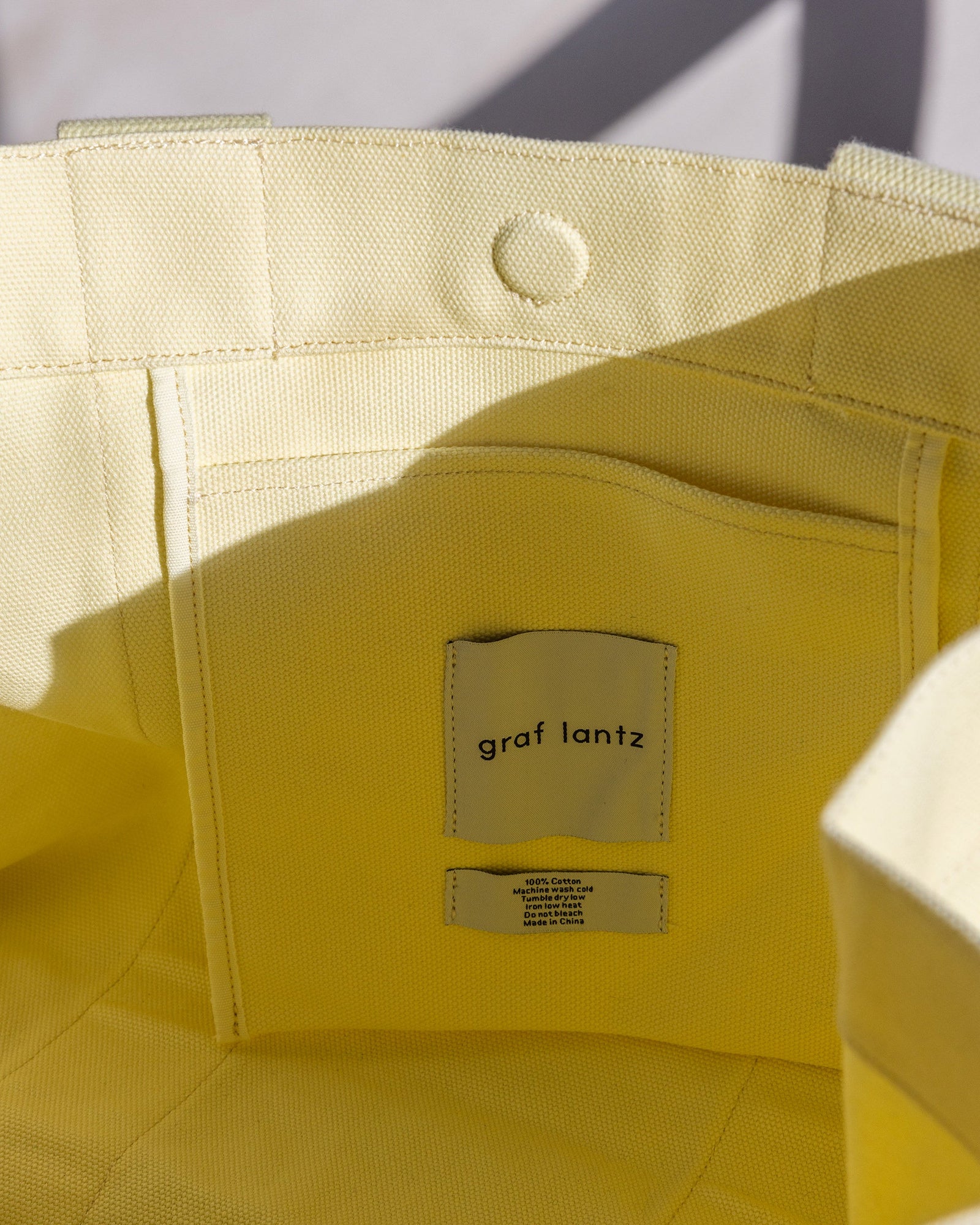 A fabric pocket with the graf lantz label is stitched inside a yellow cotton tote, showcasing its craftsmanship. The textured material catches natural light, creating subtle shadows. Ideal for beach days or casual outings, it includes a convenient magnetic closure for added security.