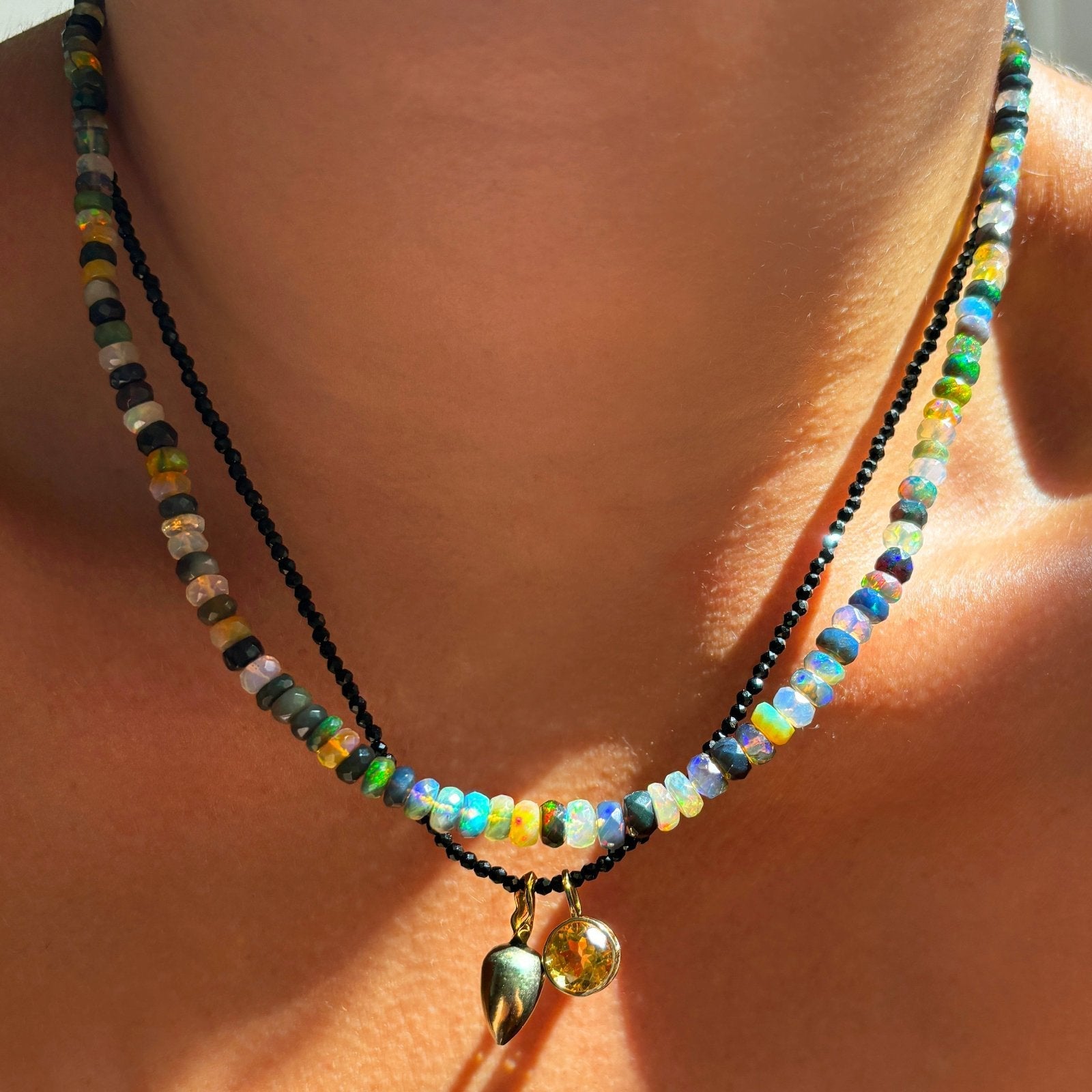 Shimmering beaded necklace made of faceted opals in shades of green, blue, purple, and white on a gold linking ovals clasp styled on a neck layered with black spinel slim gemstone and charms