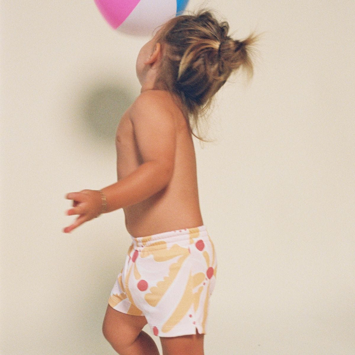 Toddler wearing white swim trunks with yellow and red palm leaf print and drawstring waistband.