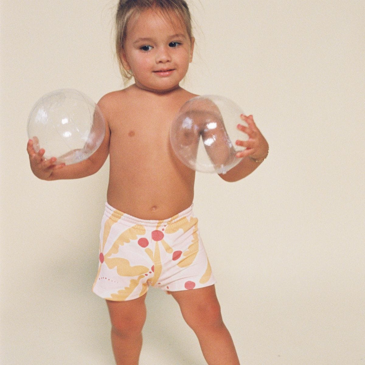Toddler wearing pink and yellow palm-patterned swim trunks with red circular details.