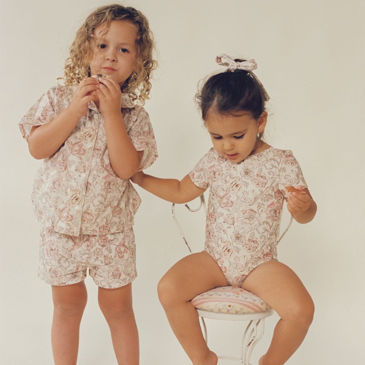 Two toddlers wearing matching cream and rose floral button-down shirts with short sleeves.